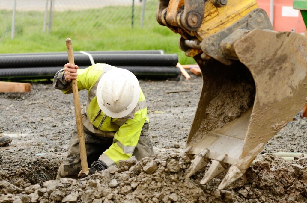 Laborer hand digging trench next to excavation bucket